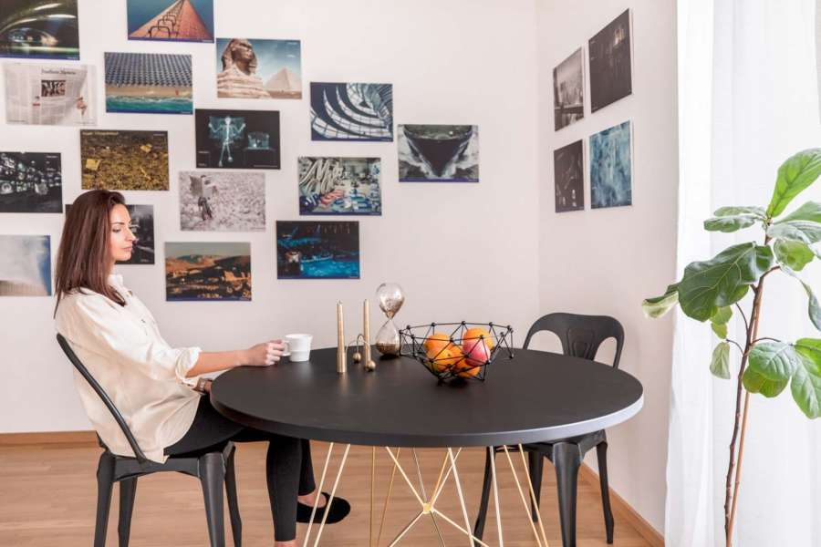 Eine Frau sitzt mit einer Tasse Kaffee an einem runden Esstisch, an der Wand hinter ihr hängen diverse Kunstdrucke