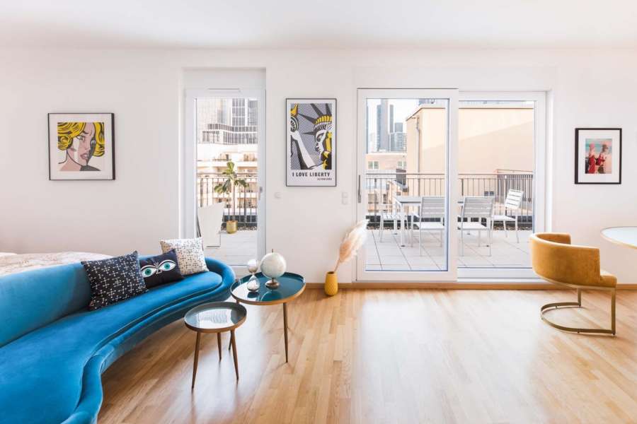 Blick aus einem modernen Apartment mit blauem Sofa in Richtung Dachterasse, im Hintergrund die Frankfurter Skyline