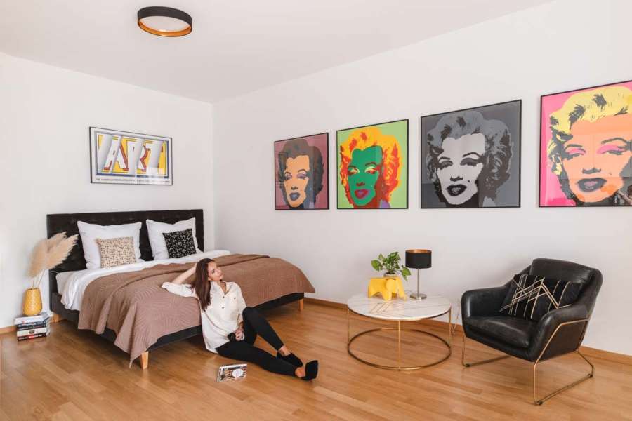 modernes Apartment mit Bett und Andy Warhol Drucken an der Wand, Sessel und Marmortisch. Eine Frau sitzt auf dem Parkettboden